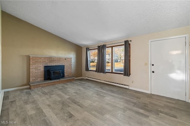 Unfurnished living room featuring vaulted ceiling, a baseboard heating unit, a wood stove, a textured ceiling, and wood finished floors