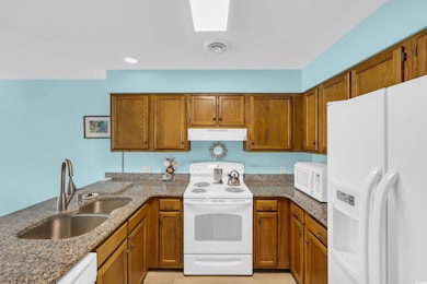 Kitchen with white appliances, under cabinet range hood, brown cabinetry, dark stone counters, and a peninsula