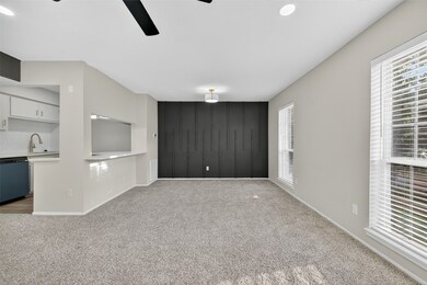 This spacious room features neutral-toned walls and plush carpeting, offering a cozy atmosphere. Large windows provide ample natural light, and a sleek black accent wall adds a modern touch. The open layout includes a view into the adjacent kitchen, enhancing connectivity.