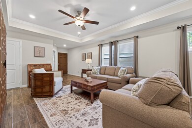 This inviting living room features a cozy seating area with plush sofas and a stylish armchair. It has warm tile flooring, a central coffee table, and soft lighting from a ceiling fan and table lamp. Large windows with curtains provide ample natural light. (Furniture available for purchase)