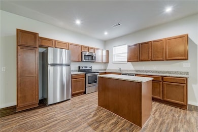 Kitchen featuring appliances with stainless steel finishes, brown cabinetry, light stone countertops, light wood-type flooring, and recessed lighting