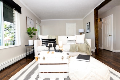 Living area featuring wood finished floors, crown molding, and baseboard heating. Home is now vacant - these photos are provided to envision the room decorated.