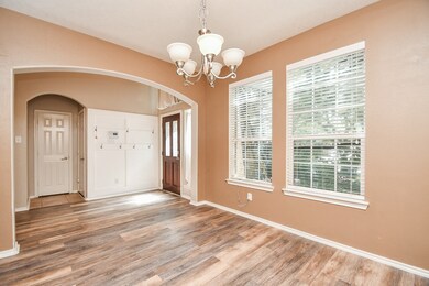 This is a bright, open room featuring wood-look flooring, a warm beige color scheme, two large windows with blinds providing natural light, and an archway leading to an adjacent space. There's a chandelier for lighting, a coat rack on the wall, and a view of the front door, suggesting this is a front entry or living area.