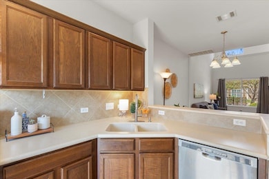 Kitchen with stainless steel dishwasher, light countertops, backsplash, brown cabinetry, and decorative light fixtures
