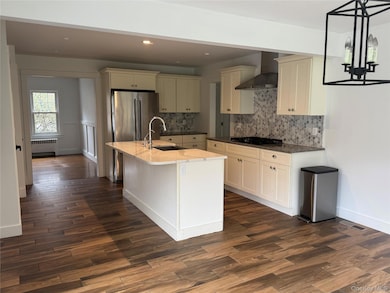 Kitchen featuring wall chimney range hood, dark stone countertops, a center island with sink, recessed lighting, and radiator heating unit