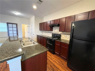 Kitchen with black appliances, tasteful backsplash, dark wood-style flooring, an island with sink, and under cabinet range hood