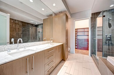 This photo showcases a modern bathroom featuring a double vanity with marble countertops, sleek wood cabinetry, and a large mirror. The space includes a glass-enclosed shower with contemporary fixtures and a doorway leading to a spacious walk-in closet with built-in shelving. The neutral color palette and tile flooring enhance the room's elegant and clean design.