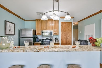 This kitchen features modern stainless steel appliances, light wooden cabinetry, and a granite countertop with a breakfast bar. Soft blue walls and warm crown molding create a cozy atmosphere, enhanced by stylish pendant lighting.