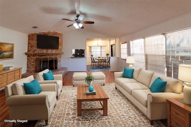This virtually staged living room features a cozy brick fireplace, large windows with blinds for natural light, and a ceiling fan. The space is decorated with neutral-toned furniture accented by blue throw pillows, creating a welcoming and comfortable atmosphere.