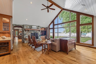 The living room impresses with a 15-foot vaulted ceiling and floor-to-ceiling picture windows that frame unobstructed lake views. Rustic white oak flooring, a stone-surround gas fireplace, and a ceiling fan create an inviting yet refined atmosphere.