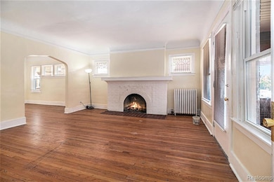 Unfurnished living room with arched walkways, hardwood / wood-style floors, healthy amount of natural light, radiator, and crown molding