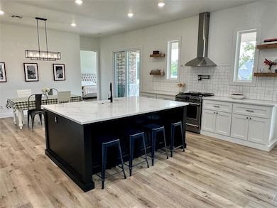 Kitchen with dark cabinets, open shelves, gas range, wall chimney range hood, and light stone countertops