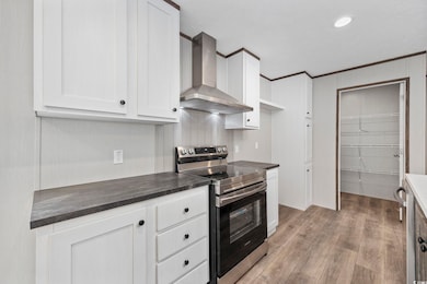 Kitchen with stainless steel range with electric cooktop, wall chimney range hood, dark countertops, white cabinetry, and light wood-style flooring