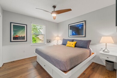 This bedroom features a modern design with hardwood floors, a ceiling fan, and ample natural light from a large window. The room is elegantly decorated with framed artwork and cozy lighting, creating a welcoming atmosphere.