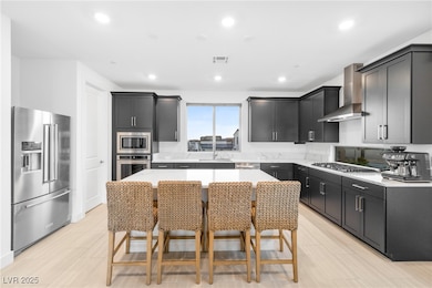 Kitchen featuring dark cabinets, stainless steel appliances, light stone counters, wall chimney range hood, and recessed lighting
