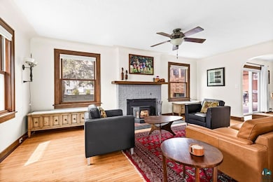 Living area featuring light wood-style floors, a ceiling fan, arched walkways, a brick fireplace, and baseboards