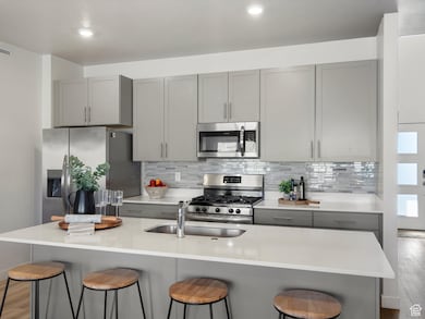 Kitchen with gray cabinetry, light wood-type flooring, appliances with stainless steel finishes, tasteful backsplash, and a center island with sink