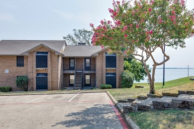 View of front facade featuring a water view, brick siding, a shingled roof, a balcony, and uncovered parking