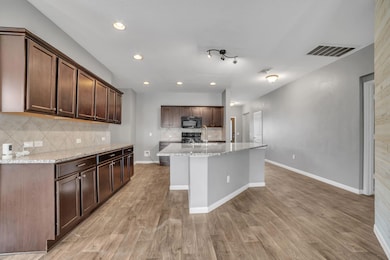 Kitchen featuring dark brown cabinets, tasteful backsplash, light stone countertops, recessed lighting, and a center island with sink