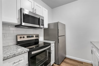 This kitchen features sleek, modern updates with stainless steel appliances, including a microwave, oven, and refrigerator. The space is enhanced by granite countertops and white cabinetry, providing a clean and contemporary look. A double-basin sink with a modern gooseneck faucet offers added convenience. The kitchen is well-lit with bright lighting, and the luxury vinyl plank adds a touch of warmth to the overall minimalist design. Ideal for those seeking a functional and stylish cooking space.