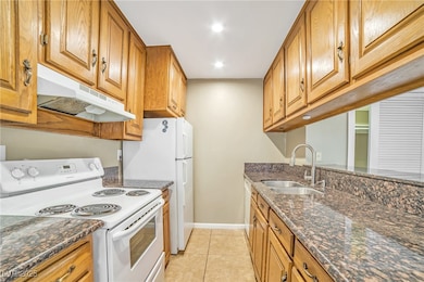Kitchen with white appliances, dark stone countertops, brown cabinetry, under cabinet range hood, and recessed lighting
