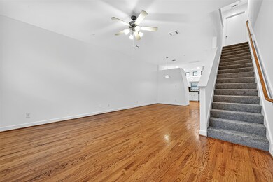 This photo showcases a bright, open living area with hardwood floors, a ceiling fan, and a staircase leading upstairs. The space flows into a kitchen area with modern lighting, creating a welcoming atmosphere.