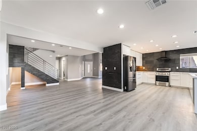 Kitchen with white cabinets, appliances with stainless steel finishes, light wood finished floors, open floor plan, and wall chimney exhaust hood