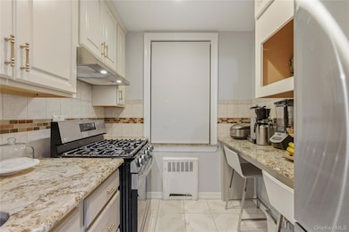 Kitchen featuring stainless steel appliances, radiator heating unit, under cabinet range hood, light stone counters, and light tile patterned flooring