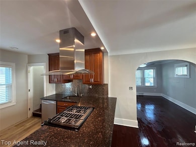 Kitchen with island exhaust hood, dark stone countertops, dark wood-type flooring, arched walkways, and backsplash