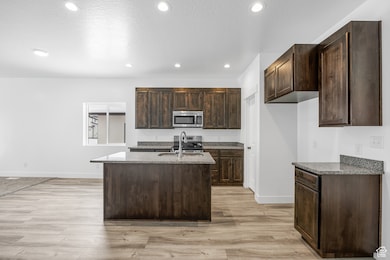 Kitchen with dark brown cabinets, dark stone counters, a kitchen island with sink, light wood-style flooring, and appliances with stainless steel finishes
