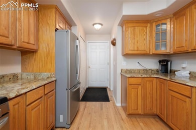 Kitchen with freestanding refrigerator, light wood finished floors, glass insert cabinets, light stone countertops, and brown cabinets