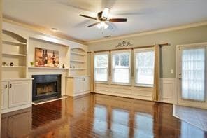 Unfurnished living room featuring crown molding, a ceiling fan, built in shelves, a fireplace, and dark wood-style flooring