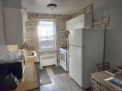 Kitchen featuring brick wall, white appliances, white cabinetry, radiator heating unit, and light countertops