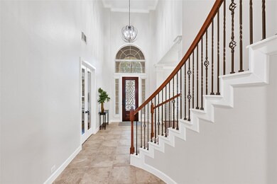 This elegant foyer features a grand curved staircase with wrought iron railings, high ceilings, and a stylish light fixture. The space is bright and welcoming, with a decorative glass front door and large tile flooring.