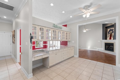 Kitchen featuring ornamental molding, light tile patterned floors, glass insert cabinets, a ceiling fan, and light stone countertops