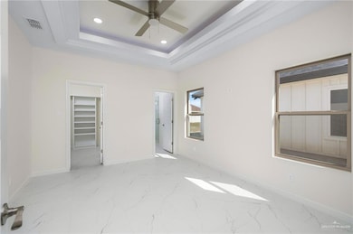 Unfurnished bedroom with a walk in closet, a raised ceiling, light marble finish floors, a ceiling fan, and recessed lighting