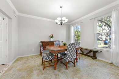 Off of the right side of the foyer, a wide entrance leads into this sunlit formal dining room with neutral grey paint, crisp white crown molding, tile floors, a 6-light brushed nickel chandelier, and a pair of front-facing windows with gauzy draperies.