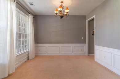 Empty room with light colored carpet, a chandelier, wainscoting, and a decorative wall