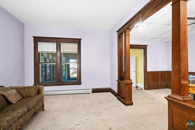 Carpeted living area with ornate columns, baseboard heating, wainscoting, and wooden walls