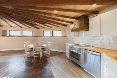 Kitchen featuring butcher block counters, custom exhaust hood, white cabinets, stainless steel appliances, and a wooden ceiling with exposed beams