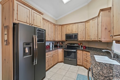 Kitchen with light brown cabinets, appliances with stainless steel finishes, crown molding, vaulted ceiling, and dark stone countertops