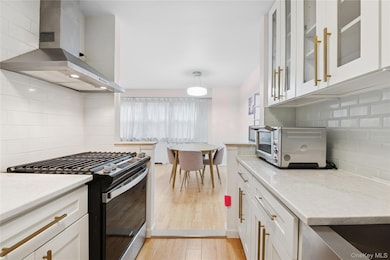 Kitchen with stainless steel range with gas stovetop, wall chimney exhaust hood, backsplash, light stone countertops, and white cabinetry