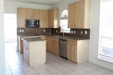 Kitchen with light brown cabinetry, appliances with stainless steel finishes, backsplash, and light stone countertops