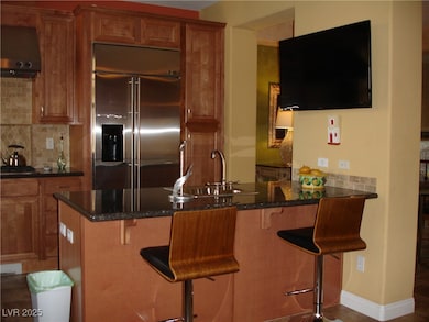 Kitchen featuring stainless steel built in refrigerator, exhaust hood, brown cabinetry, tasteful backsplash, and dark stone countertops