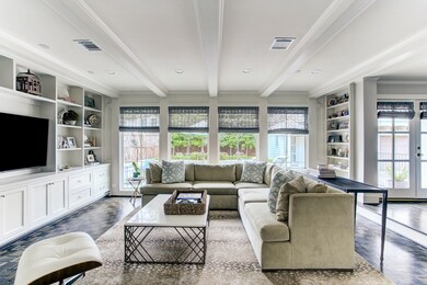 The welcoming family room overlooks the immaculate backyard. A wall of floor-to-ceiling windows look out onto the back patio and give stunning views of the sparkling pool. Notice the exposed beams and parquet hardwood floors, one of the many pristine touches of this special residence.