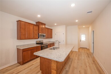 Kitchen featuring brown cabinets, appliances with stainless steel finishes, light wood finished floors, light stone counters, and a center island with sink