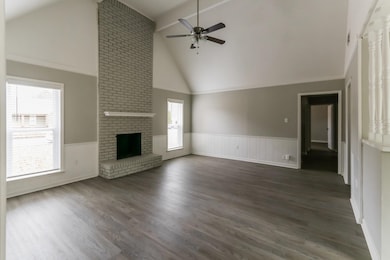 Unfurnished living room with high vaulted ceiling, wainscoting, dark wood-type flooring, a brick fireplace, and a ceiling fan