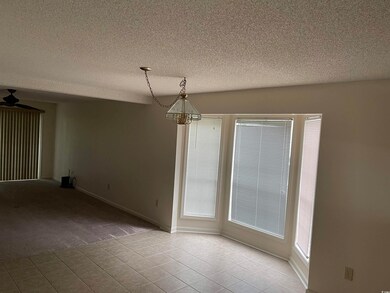 Carpeted living room with a textured ceiling, a chandelier, tiled dining area with bay windows.