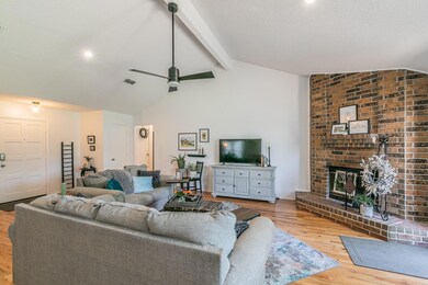 Living room featuring light hardwood / wood-style flooring, a brick fireplace, vaulted ceiling with beams, brick wall, and ceiling fan