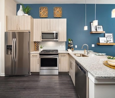Kitchen featuring stainless steel appliances, light stone counters, a peninsula, light brown cabinetry, and dark wood-style floors